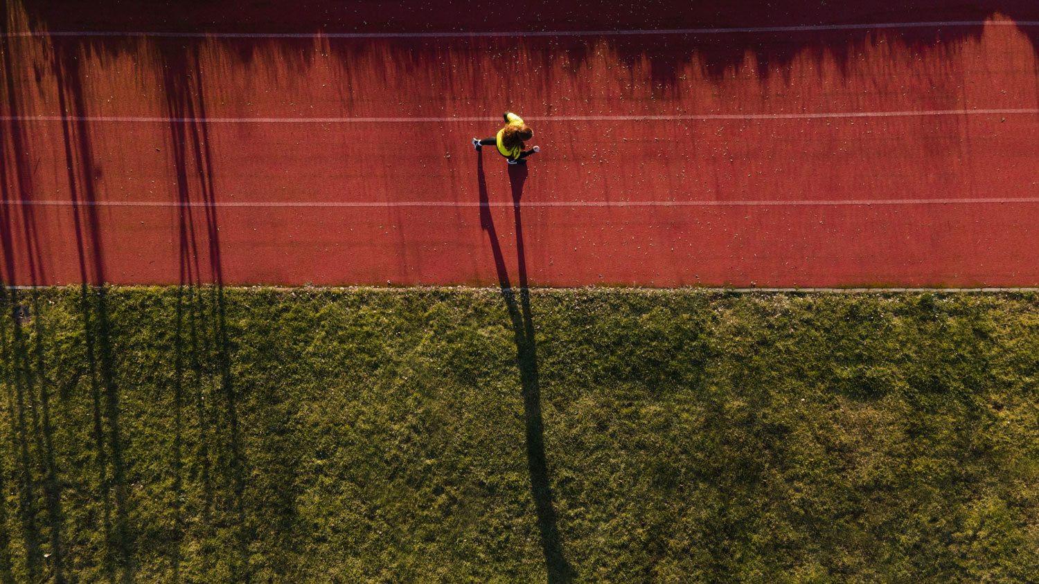 Vue d'en haut d'une personne courant sur une piste d'athlétisme et son ombre projettés sur le sol à côté